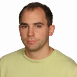 Portret mężczyzny w jasnozielonym swetrze na białym tle, z delikatnym uśmiechem i widocznym logo na ubraniu. Ujęcie od przodu, skupiające się na twarzy.