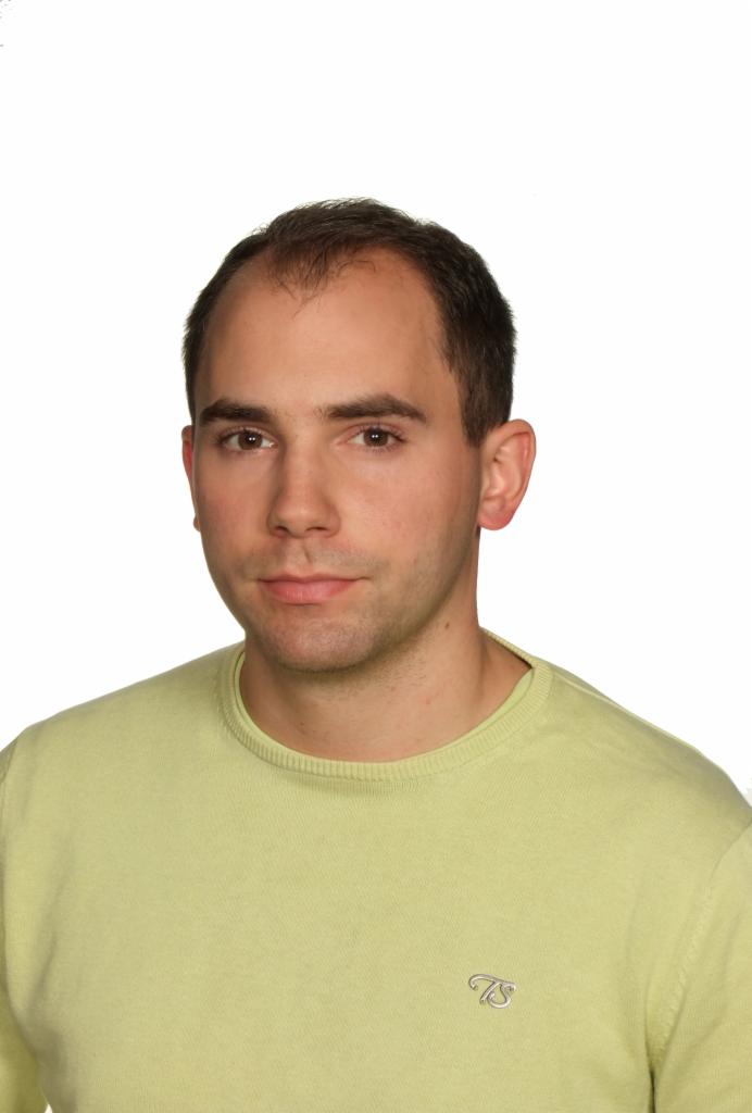 Portret mężczyzny w jasnozielonym swetrze na białym tle, z delikatnym uśmiechem i widocznym logo na ubraniu. Ujęcie od przodu, skupiające się na twarzy.