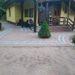 Kaskadowa ścieżka z kostki brukowej w odcieniach czerwieni i szarości prowadząca do domu z drewnianym gankiem, widoczny piasek przed ułożoną kostką i niebieska zmiotka w prawym dolnym rogu.