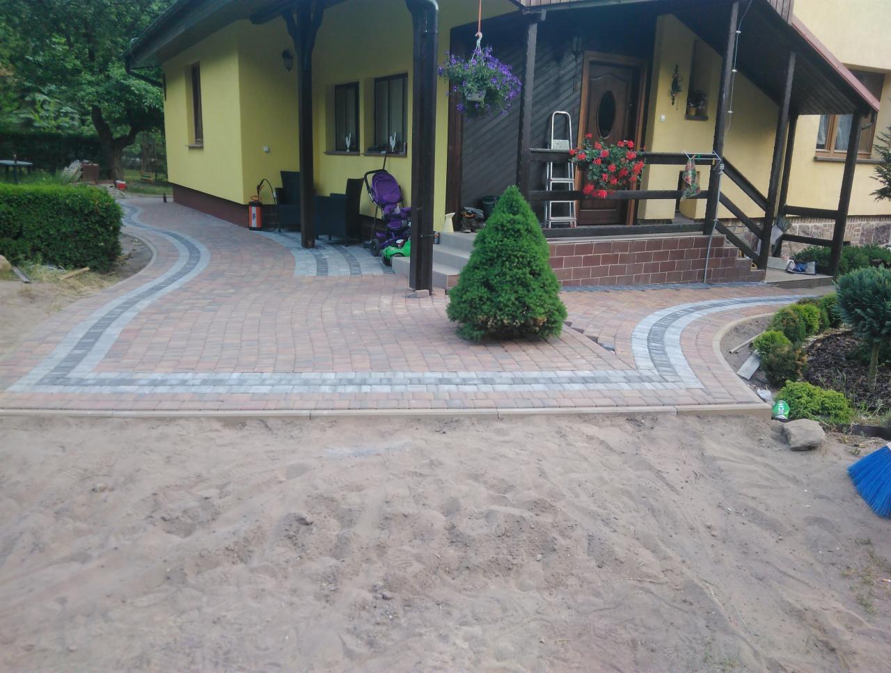 Kaskadowa ścieżka z kostki brukowej w odcieniach czerwieni i szarości prowadząca do domu z drewnianym gankiem, widoczny piasek przed ułożoną kostką i niebieska zmiotka w prawym dolnym rogu.