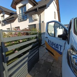 KLIMAT Adam Kordek - Biały budynek jednorodzinny z zainstalowaną jednostką zewnętrzną klimatyzacji, widoczny zza otwartej furtki i samochodu firmowego z napisem 'KLIMAT'.