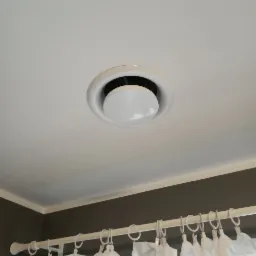 Okrągły, biały nawiewnik klimatyzacji sufitowej w pomieszczeniu z szarą ścianą i białymi zasłonami.