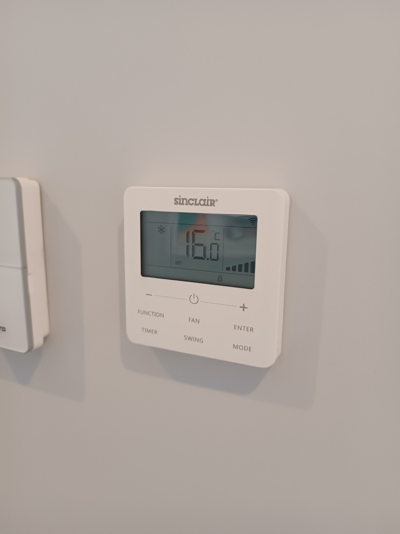 Biały, elektroniczny panel sterowania klimatyzacją marki Sinclair zamontowany na ścianie, wyświetlający temperaturę 16 stopni Celsjusza, z widocznym panelem sterowania obok.
