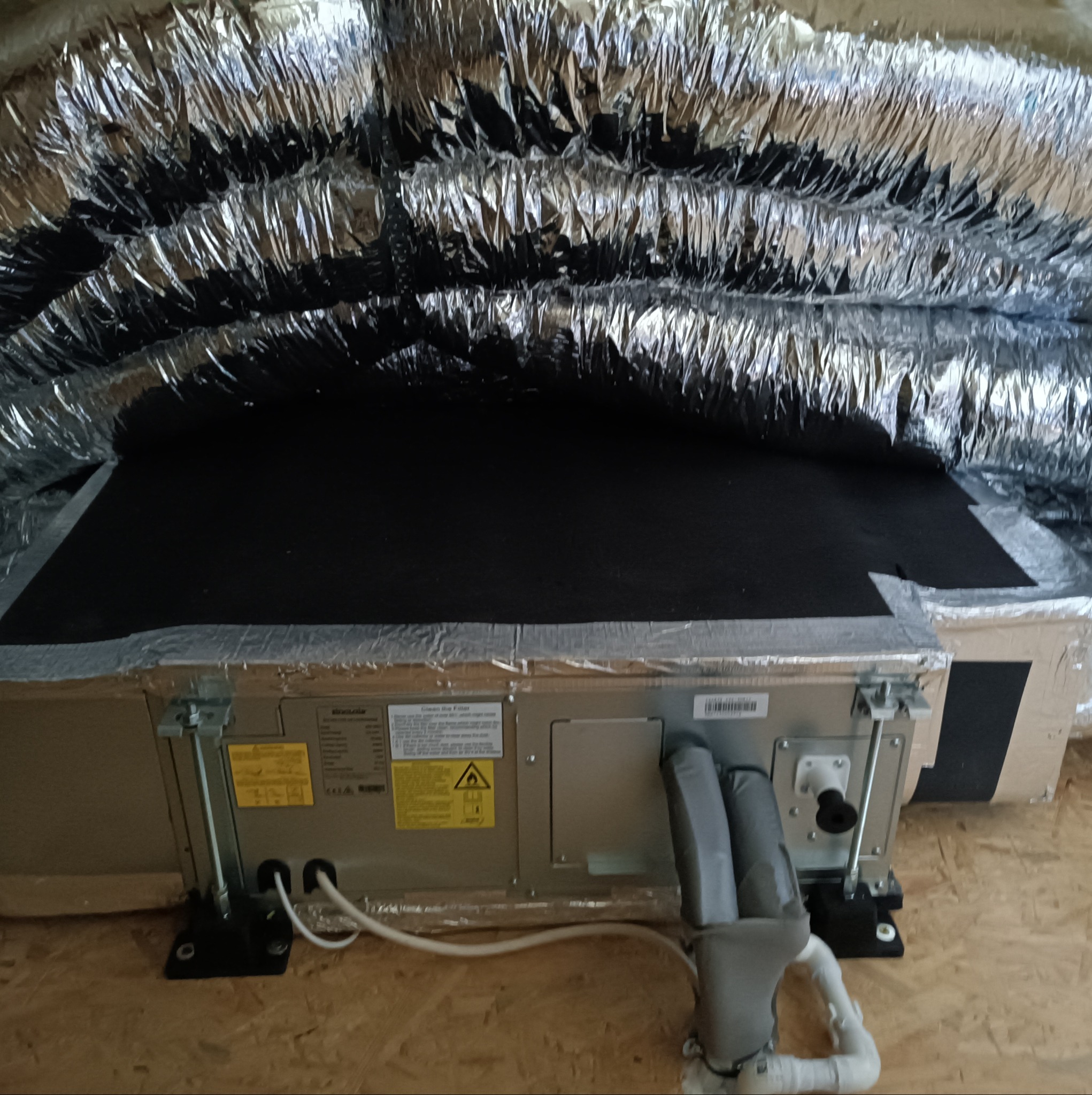 Widok z dołu na zamontowaną jednostkę wewnętrzną klimatyzacji kanałowej z widocznymi przewodami izolowanymi termicznie i elementami mocującymi, umieszczoną na drewnianej powierzchni.