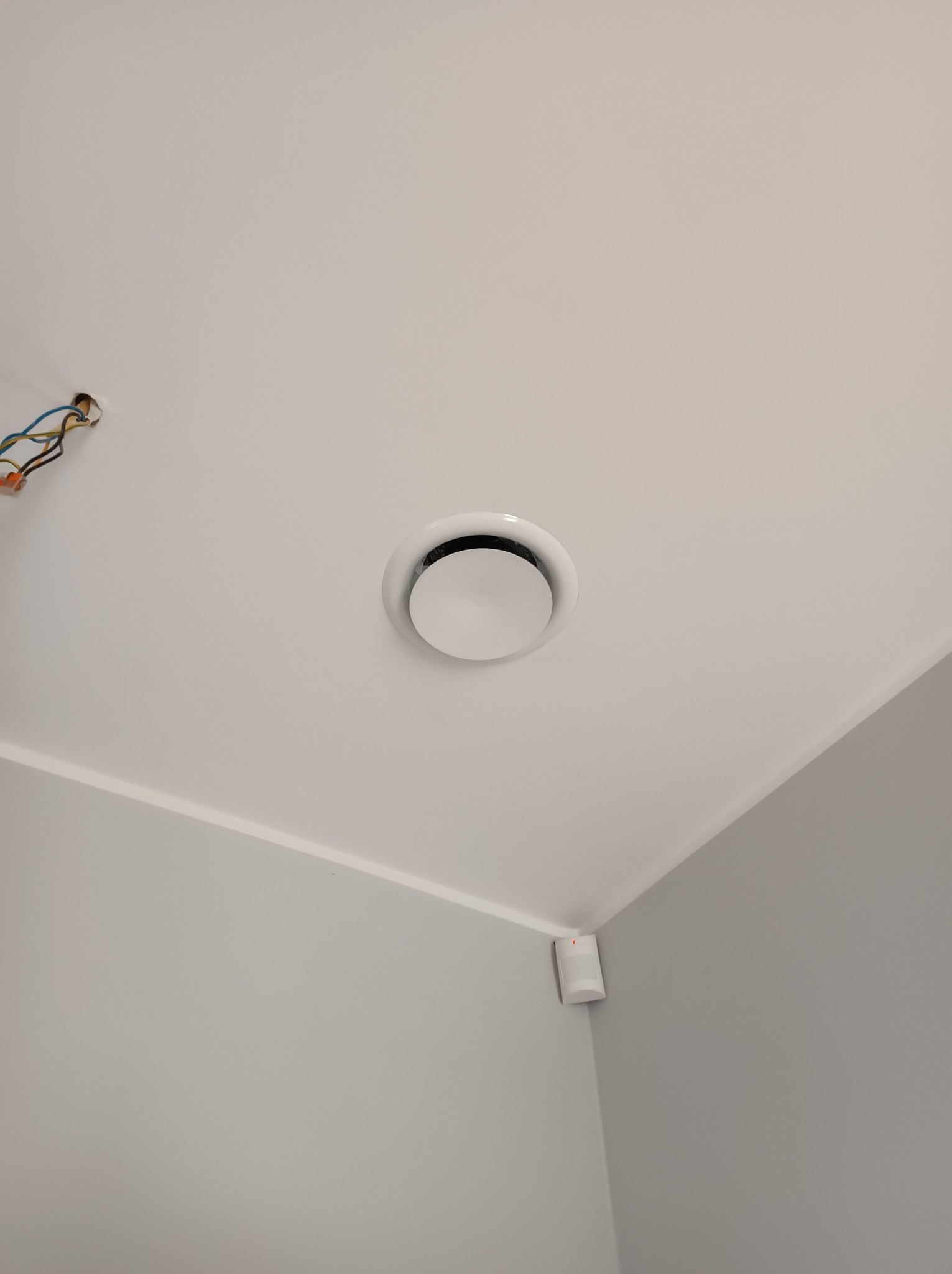 Okrągły nawiewnik sufitowy w białym pomieszczeniu, widoczne wystające przewody elektryczne w rogu sufitu.