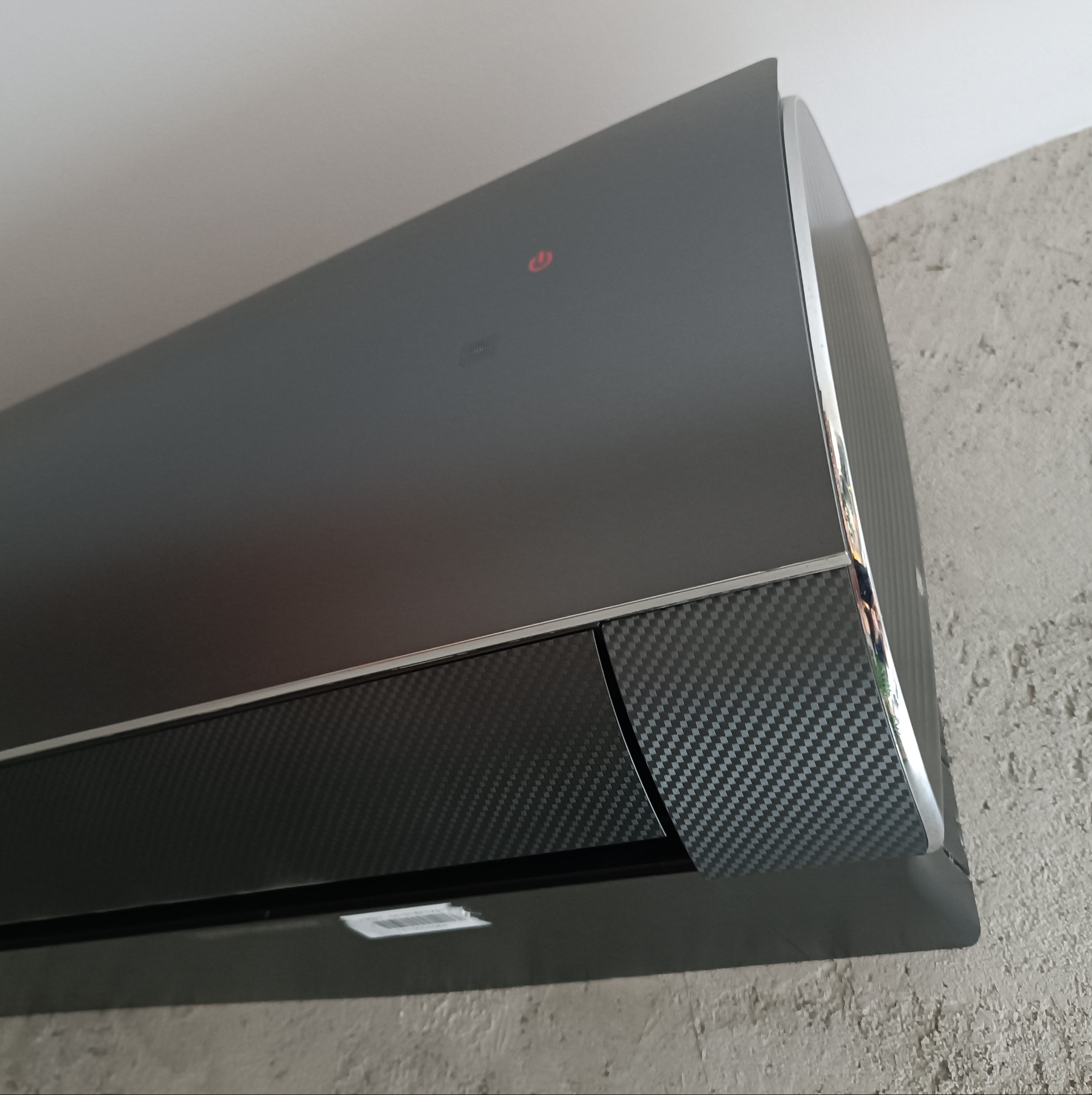 Czarny klimatyzator ścienny z panelem w stylu karbonu, z widoczną czerwoną diodą zasilania, zamontowany na szarej, chropowatej ścianie.