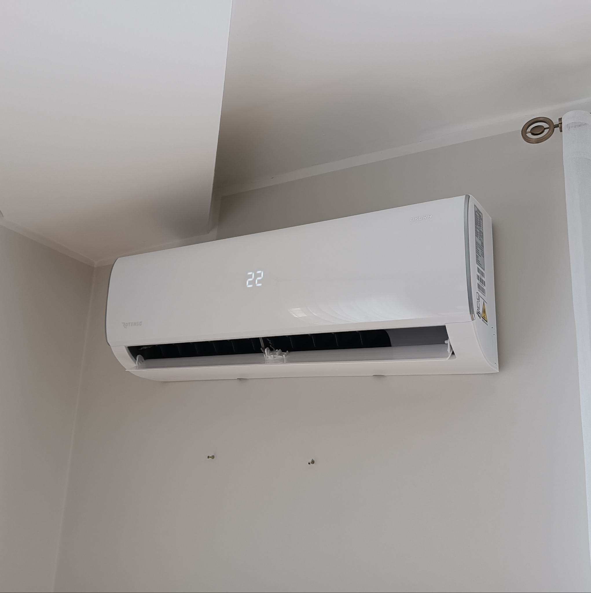 Biały klimatyzator marki Rotenso, model Ukura, zamontowany na szarej ścianie w pomieszczeniu. Na wyświetlaczu widoczna temperatura 22 stopnie Celsjusza. Widoczne ślady po poprzednim montażu.