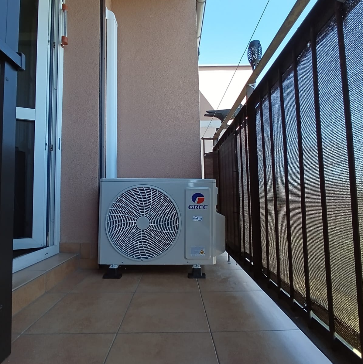 Zamontowana jednostka zewnętrzna klimatyzacji Gree na balkonie z brązowymi płytkami i ciemną balustradą.