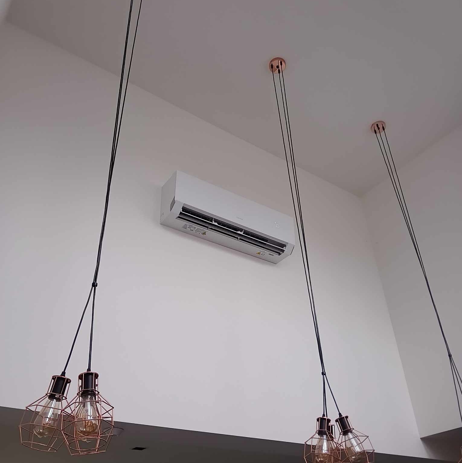 Nowoczesny klimatyzator ścienny w białym wnętrzu, widoczny spod kątem, z dekoracyjnymi lampami wiszącymi w stylu industrialnym z miedzianymi elementami.