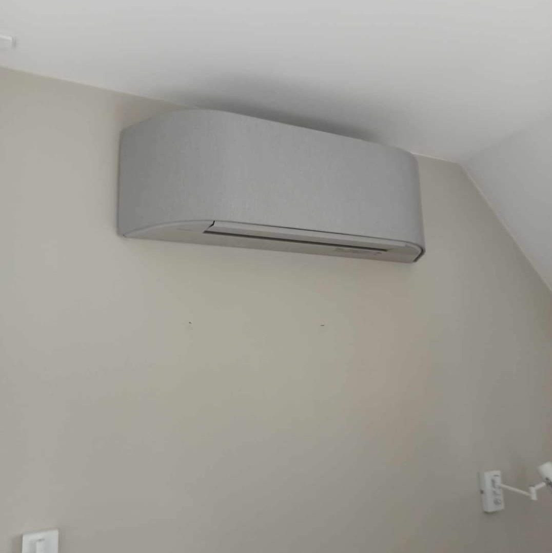 Szary klimatyzator ścienny o zaokrąglonym kształcie, zamontowany w rogu pomieszczenia, widoczne gniazdko elektryczne na ścianie.