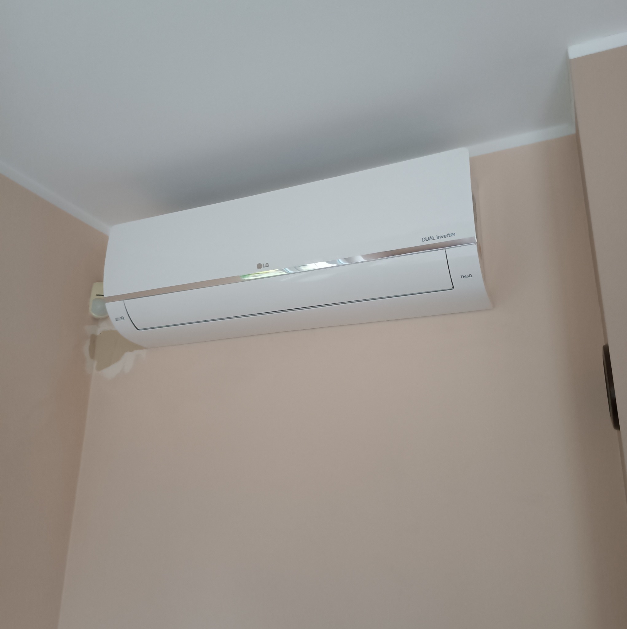 Biała jednostka wewnętrzna klimatyzacji LG Dual Inverter zamontowana na beżowej ścianie w pomieszczeniu.