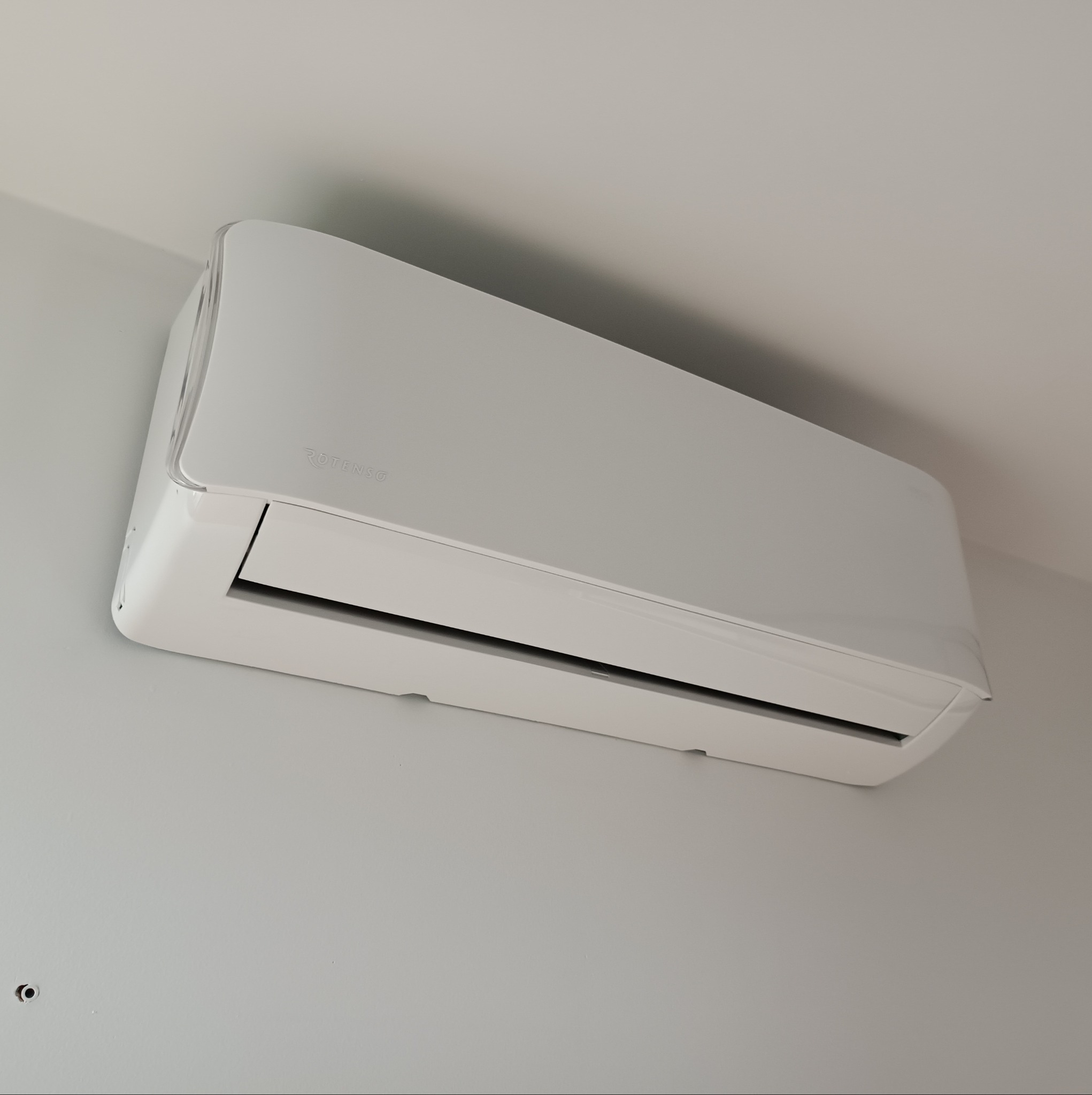 Biały klimatyzator Rotenso zamontowany na jasnej ścianie, widoczne delikatne odbicia światła na obudowie.