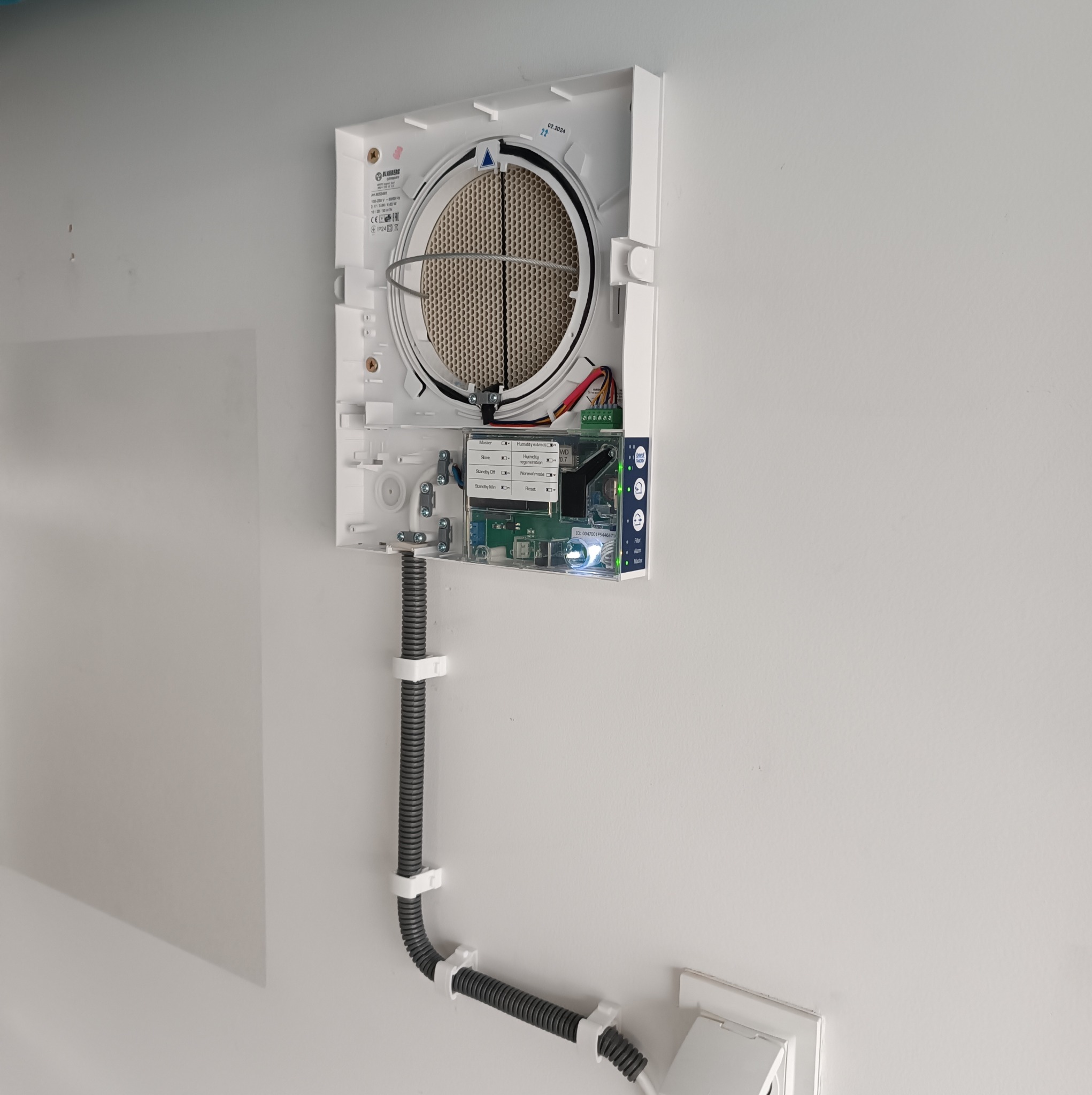 Centrala wentylacyjna z widocznym panelem sterowania, zamontowana na ścianie, z przewodami w elastycznej rurze przymocowanej klamrami.