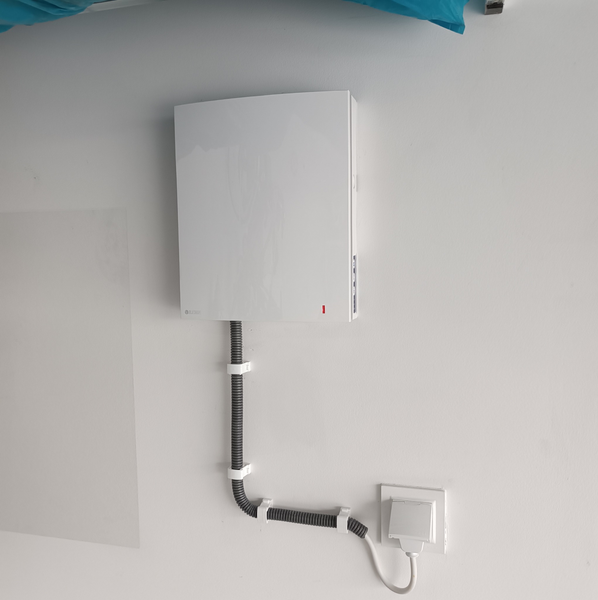 Biała jednostka wewnętrzna klimatyzacji z podłączonym szarym przewodem w peszlu, zamocowanym do ściany za pomocą białych uchwytów, obok gniazdka elektrycznego z wtyczką.