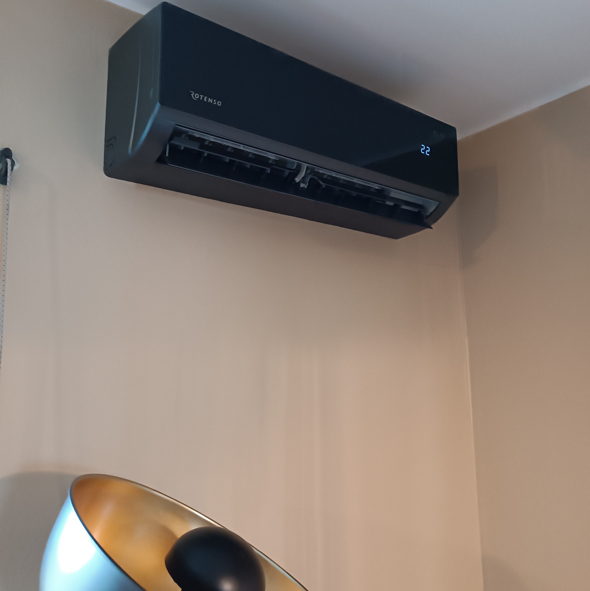 Czarna jednostka klimatyzacyjna marki Rotenso zamontowana na ścianie w pomieszczeniu, z wyświetlaczem temperatury ustawionym na 22 stopnie Celsjusza, widoczna lampa stojąca z białym kloszem...