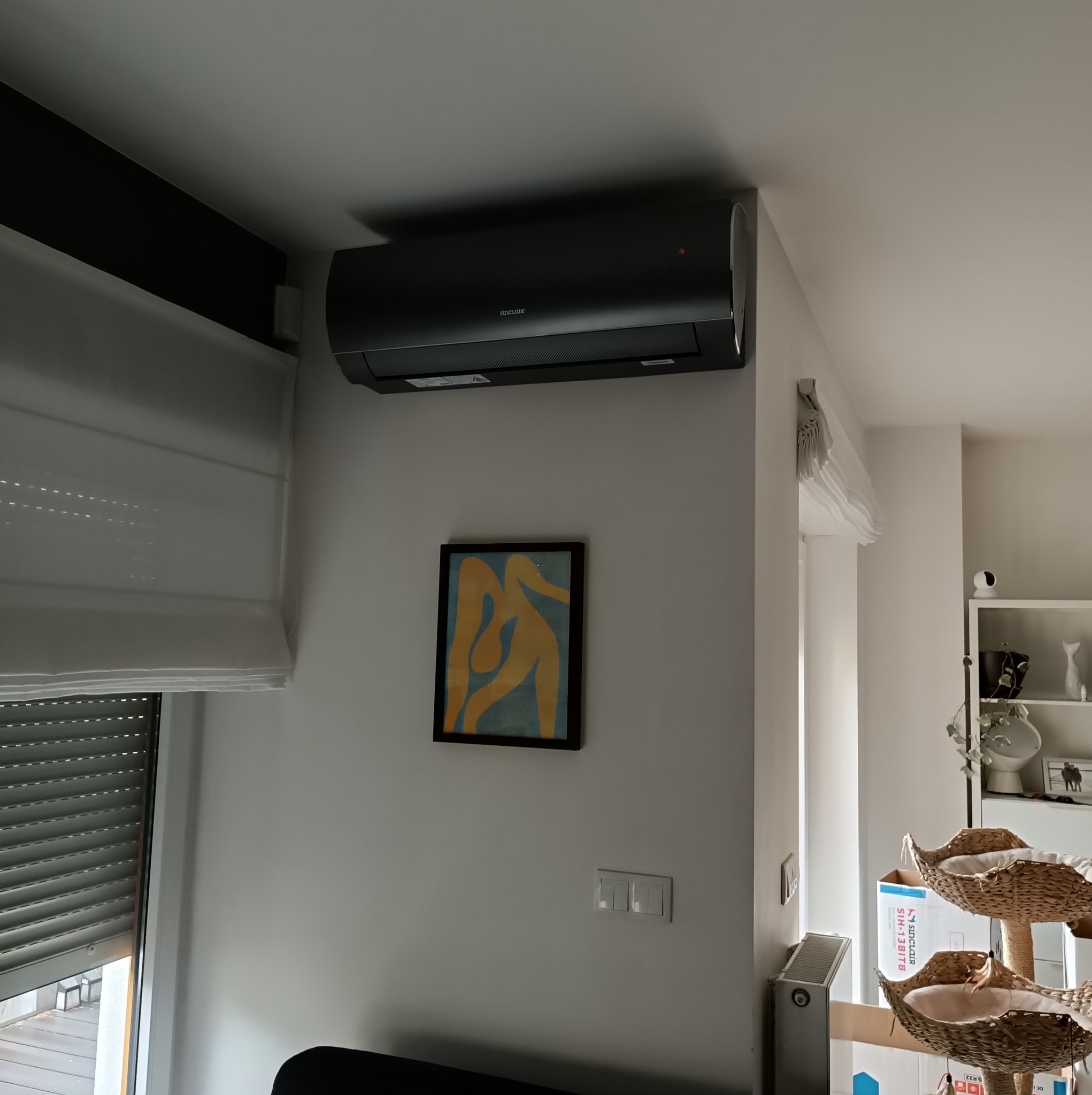 Czarny klimatyzator Sinclair zamontowany na ścianie w mieszkaniu, pod nim wisi obraz w ramie, a obok okno z roletami.