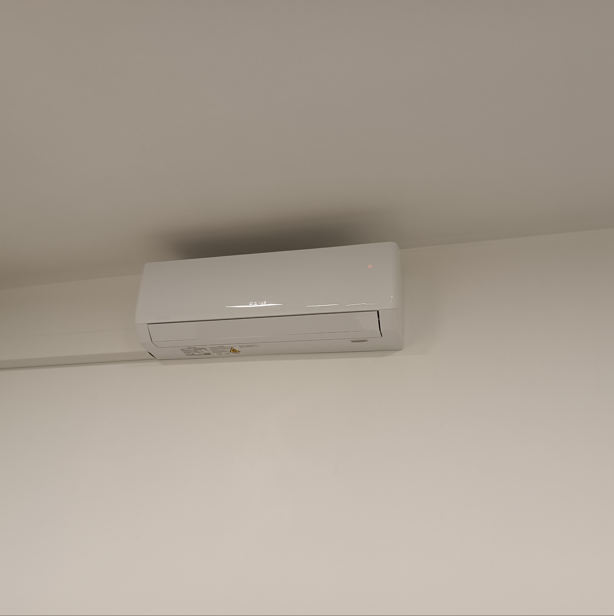 Biała jednostka klimatyzacji zamontowana na jasnej ścianie, widoczna niewielka czerwona dioda sygnalizacyjna, w tle biały sufit.