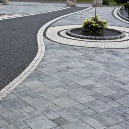 Nowoczesny podjazd z szarej kostki brukowej ułożonej we wzór, z elementami dekoracyjnymi z jasnej kostki i ciemnego żwiru, zakończony okrągłym klombem z małym drzewkiem.