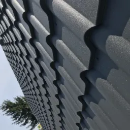 Szary dach z blachy trapezowej z widoczną rynną i fragmentem drzewa na tle nieba. Ujęcie z dołu, ukazujące regularny wzór pokrycia.