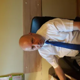 Mężczyzna w białej koszuli i niebieskim krawacie siedzi na krześle biurowym, widoczny kalendarz na ścianie.