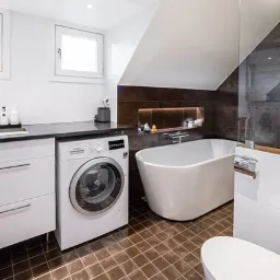 Nowoczesna łazienka na poddaszu z brązowymi płytkami podłogowymi, białą wanną wolnostojącą, pralką Bosch obok białych szafek i toaletą.
