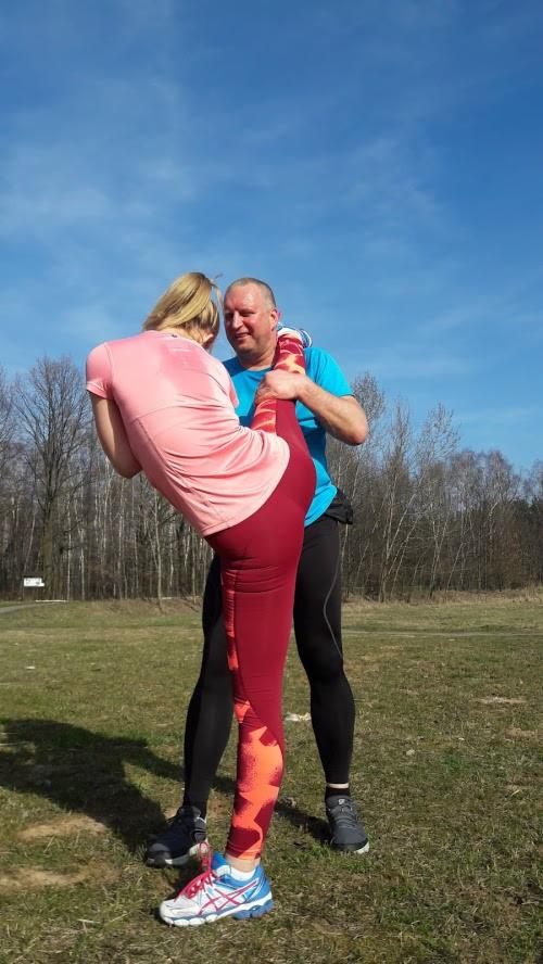 Kobieta w różowej koszulce i bordowych legginsach rozciąga się na zewnątrz, trzymając nogę uniesioną przez mężczyznę w niebieskiej koszulce na tle drzew i błękitnego nieba.