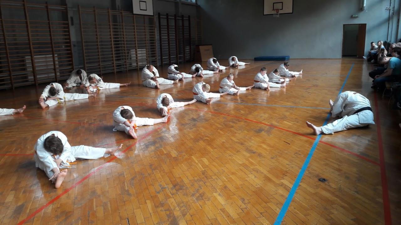 Grupa dzieci w białych kimonach rozciąga się na drewnianej podłodze sali gimnastycznej podczas treningu sportów walki, w tle widoczni obserwatorzy.