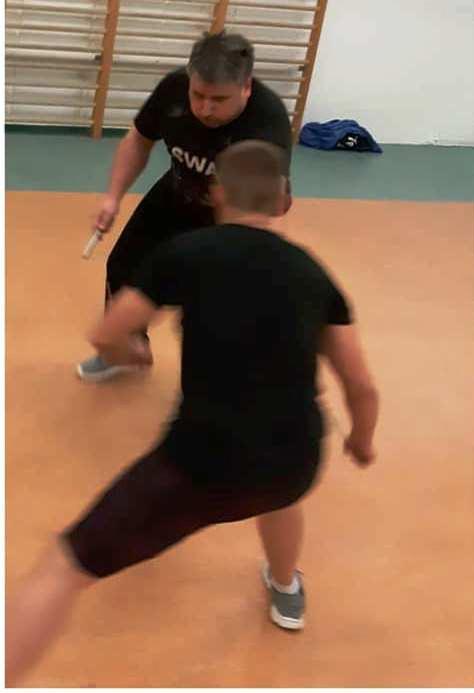 Instruktor z krótką pałką w dłoni uczy samoobrony, uczeń w czarnym stroju wykonuje kopnięcie w pomieszczeniu z drabinką gimnastyczną.