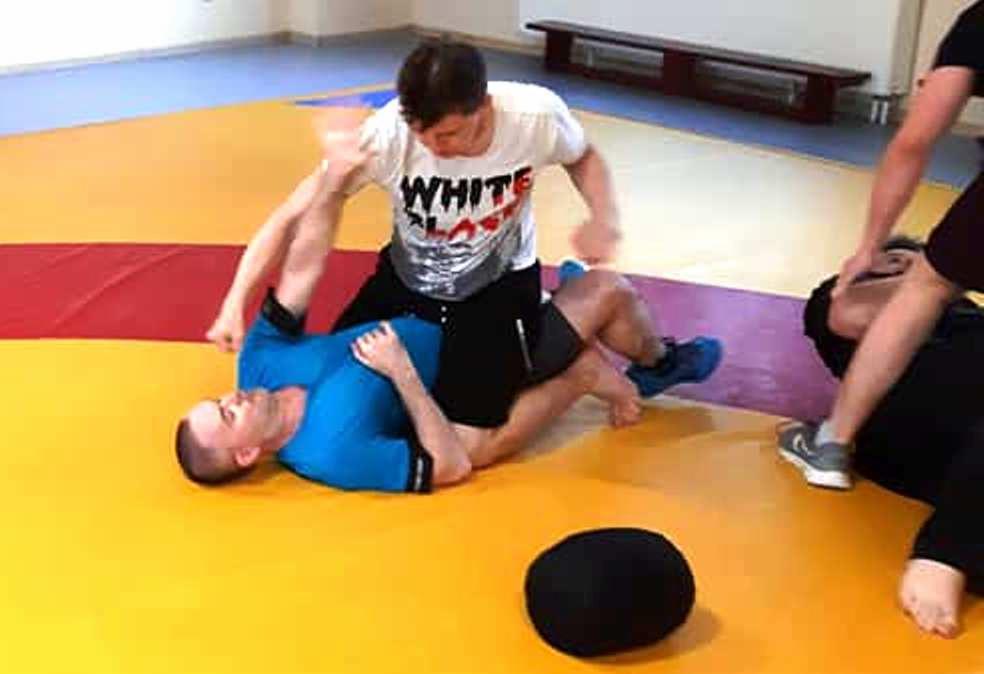 Trening sportów walki: mężczyzna w białej koszulce z napisem 'WHITE' w pozycji dominującej na przeciwniku w niebieskiej koszulce na macie w sali treningowej.