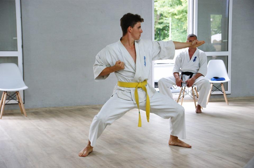 Młody mężczyzna w białym kimonie z żółtym pasem wykonuje pozycję obronną karate w jasnym pomieszczeniu treningowym, w tle siedzący mężczyzna w kimonie na krześle.