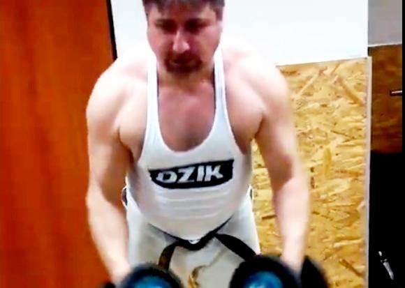 Mężczyzna w białym podkoszulku z napisem 'DZIK' ćwiczy podnoszenie ciężarów w siłowni, widok z perspektywy osoby ćwiczącej.