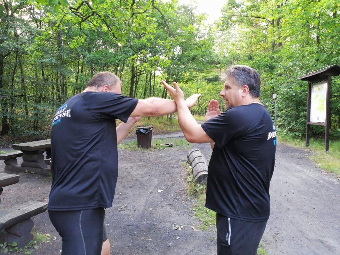Dwóch mężczyzn w czarnych koszulkach trenuje techniki samoobrony na zewnątrz, w tle widoczny las i drewniane ławki.