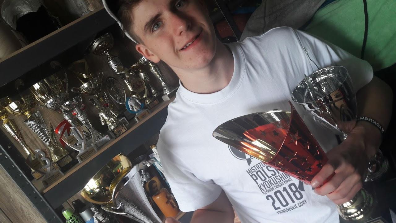 Młody mężczyzna uśmiecha się, trzymając dwa puchary, na tle półki z licznymi trofeami; na koszulce nadruk 'Mistrzostwa Polski Kyokushin 2018'.