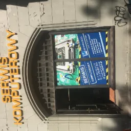 Fasada budynku z napisem 'SERWIS KOMPUTEROWY' nad wejściem, oklejona plakatami reklamującymi usługi naprawy laptopów i odzyskiwania danych, z widoczną płytą główną komputera na plakacie.