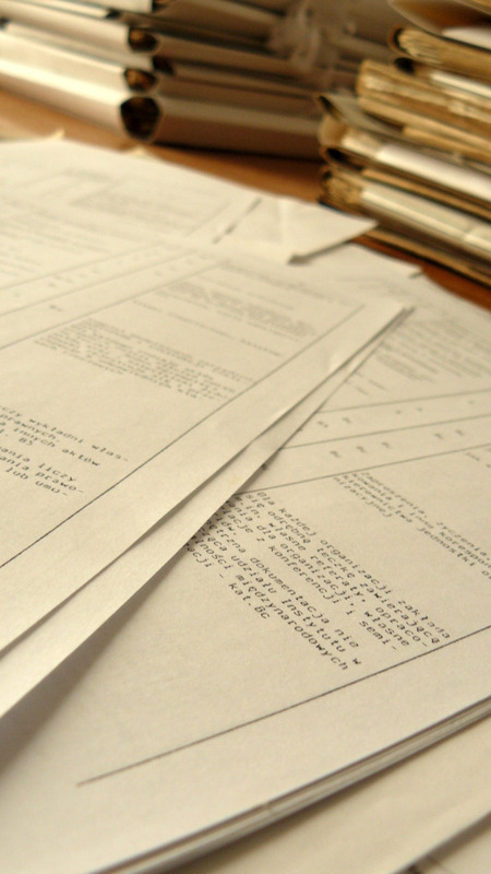 Stos dokumentów tekstowych z widocznymi fragmentami treści, ułożonych na blacie biurka obok segregatorów.