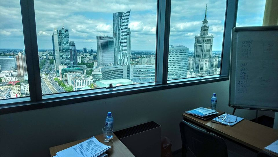 Widok z okna biurowca na panoramę Warszawy, w tle Pałac Kultury i Nauki, na biurku butelka wody, dokumenty, okulary i tablica suchościeralna z zapiskami.