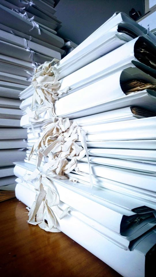 Stosy białych segregatorów z dokumentami, zabezpieczonych bawełnianymi tasiemkami, ułożone na drewnianym biurku, w tle kolejne stosy.