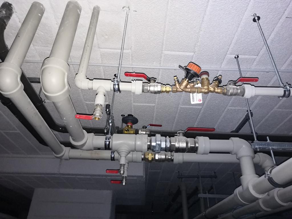 Skomplikowany układ rur instalacyjnych pod sufitem z widocznymi zaworami z czerwonymi uchwytami, obejmami mocującymi i elementami mosiężnymi.