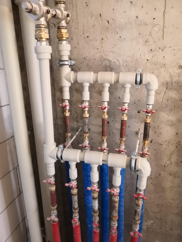 Rozdzielacz instalacji wodnej z rurami PP i zaworami kulowymi na tle surowej ściany betonowej z odręcznymi notatkami.