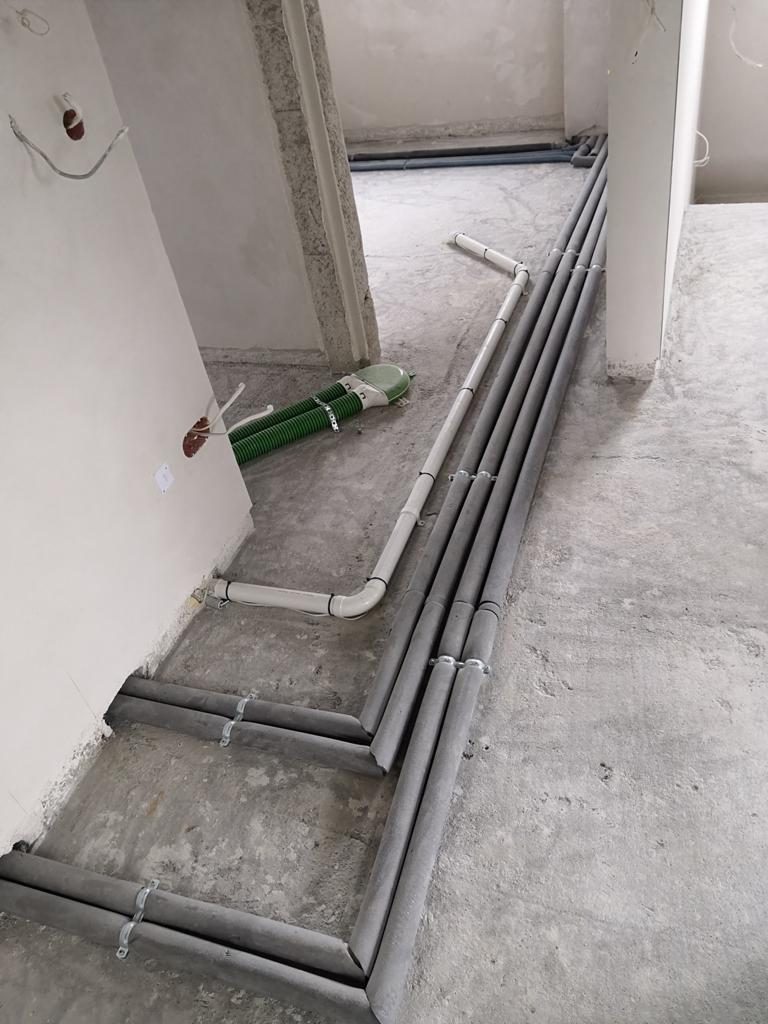 Szare rury izolowane biegnące wzdłuż podłogi w surowym stanie, przymocowane metalowymi uchwytami, obok widoczne białe rury i zielone karbowane przewody wentylacyjne.