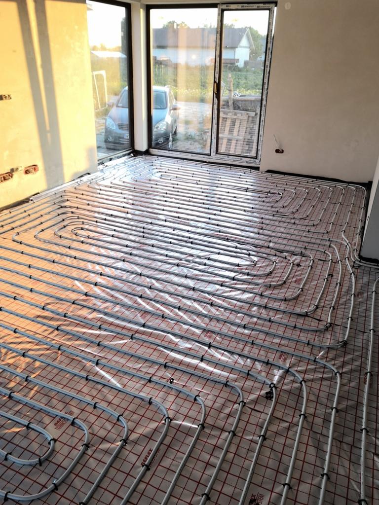 Układanie rur ogrzewania podłogowego na folii refleksyjnej z czerwoną siatką w jasnym, surowym wnętrzu z oknem na ogród i zaparkowany samochód.