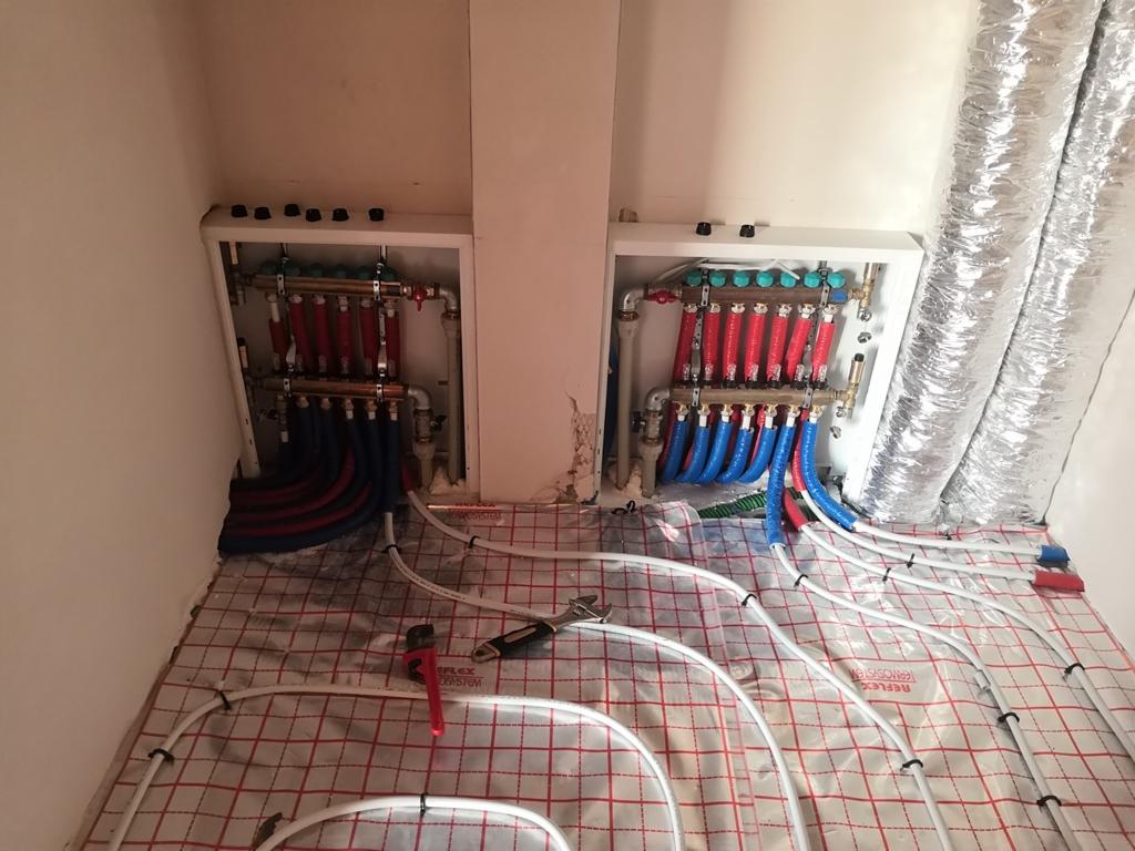 Rozdzielacze ogrzewania podłogowego w szafkach, z widocznymi rurami doprowadzającymi i odprowadzającymi wodę, ułożone na folii izolacyjnej z czerwoną siatką, klucz nastawny leżący obok rur.