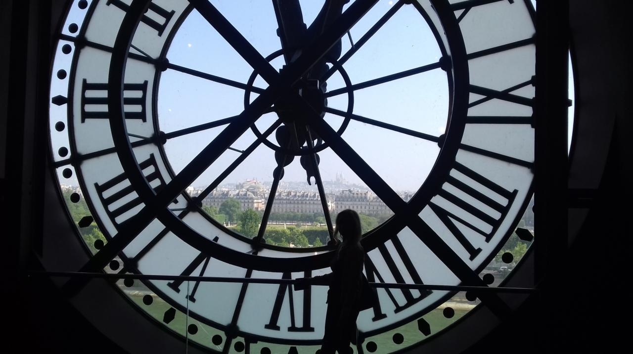 Sylwetka kobiety na tle ogromnego zegara w Musee d'Orsay z widokiem na panoramę Paryża w tle.