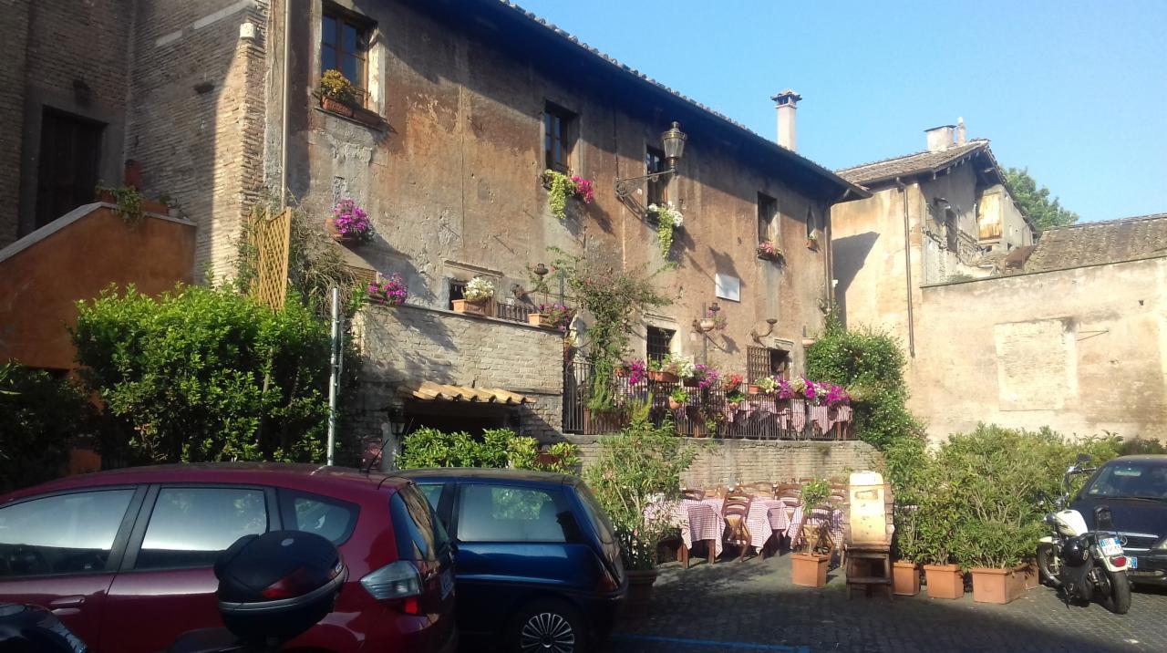 Rustykalny budynek z balkonem udekorowanym kwiatami, stoliki restauracyjne na zewnątrz z czerwono-białymi obrusami, zaparkowane samochody w tle.