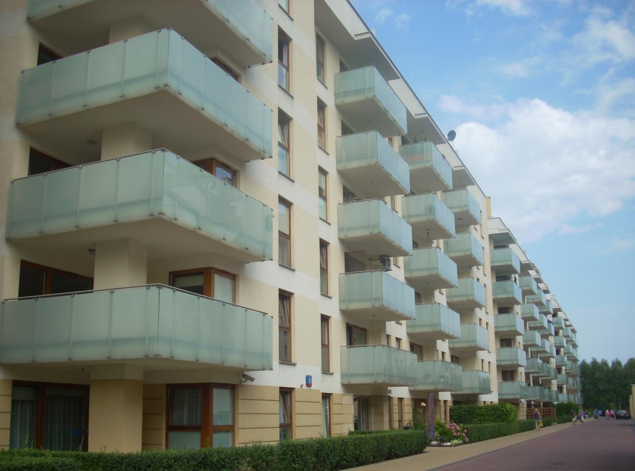 Fasada budynku wielorodzinnego z balkonami zabezpieczonymi mlecznym szkłem, widok z perspektywy chodnika z zielenią.