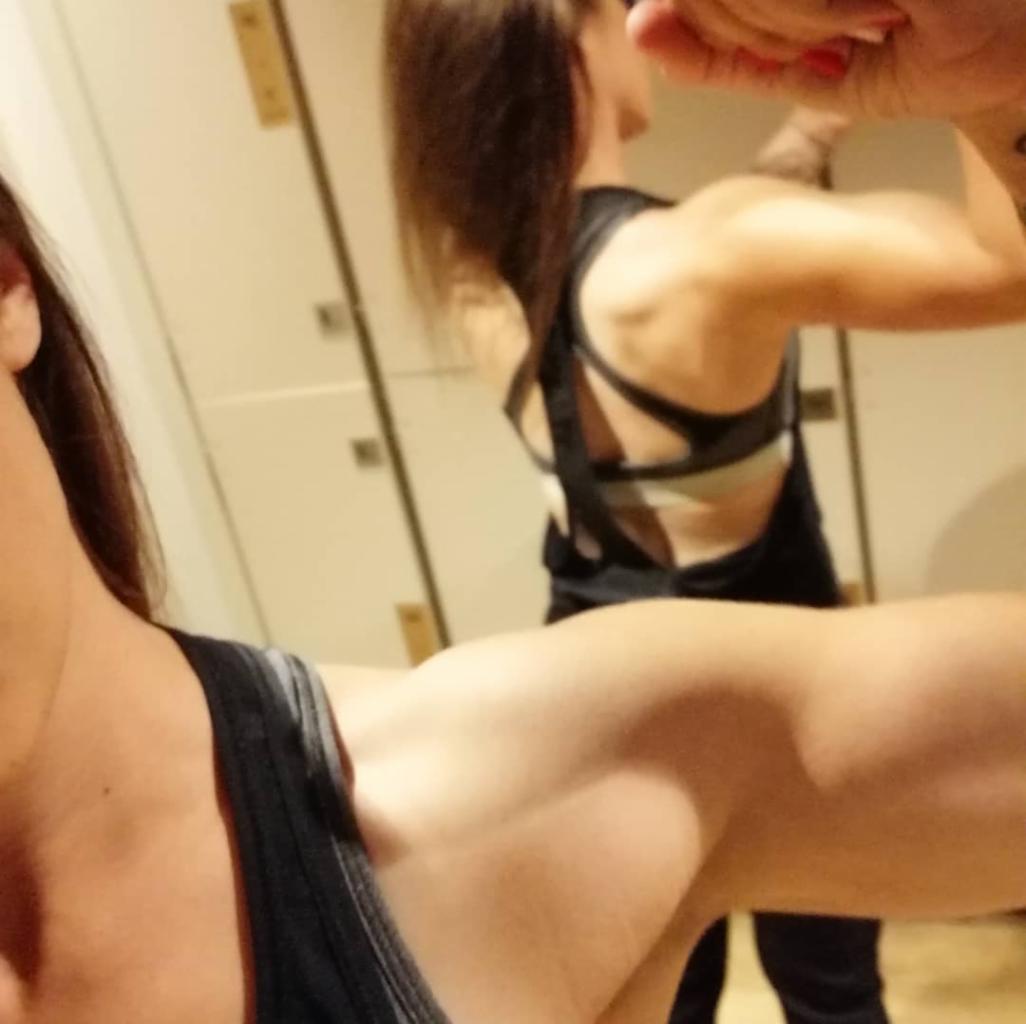 Kobieta prezentuje umięśnione ramię w lustrze, ubrana w sportowy top z paskami na plecach. Widoczny fragment drugiego ramienia i szafki w tle.