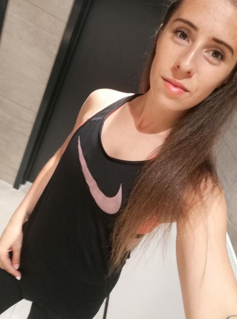 Kobieta w czarnej koszulce sportowej z różowym logo Nike, w szatni, selfie z góry, długie włosy, naturalny makijaż, widoczny fragment tatuażu na ręce.
