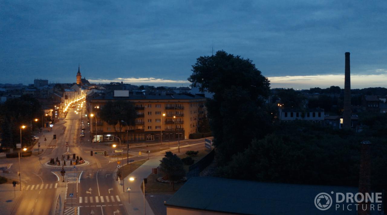 Nocny widok z drona na miasto z oświetloną ulicą, budynkami i kominem w tle, logo 'Drone Picture' w prawym dolnym rogu.