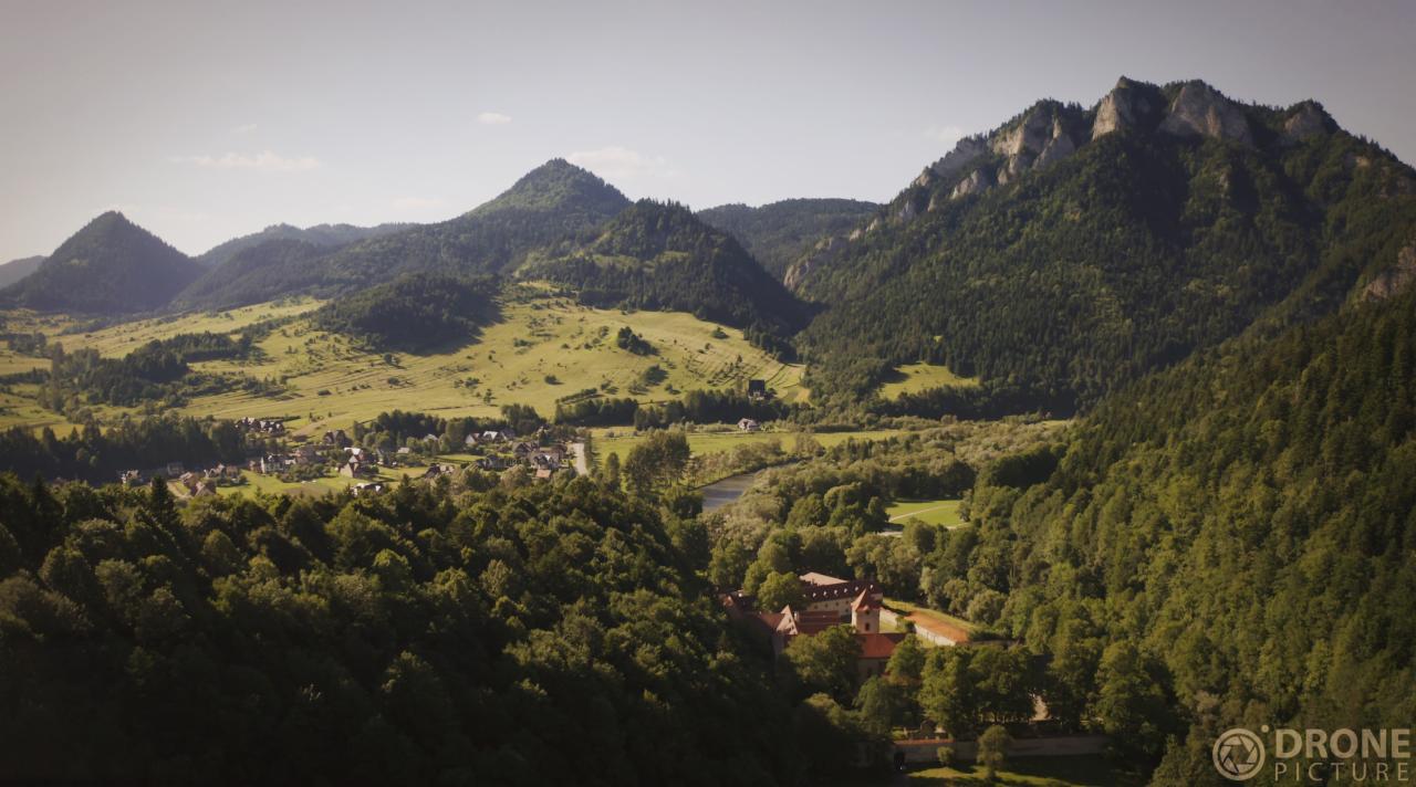 Panoramiczny widok z drona na malowniczą dolinę z rzeką, lasami i górami w tle, z widocznym kościołem w oddali i zabudowaniami w dolinie.