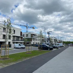 Nowe osiedle mieszkaniowe z chodnikiem wyłożonym szarą kostką brukową, świeżo posadzone drzewa zabezpieczone drewnianymi konstrukcjami, samochody zaparkowane wzdłuż drogi, w tle żurawie budowlane.